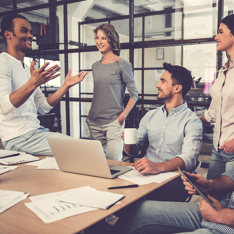 A group of coworkers sits and stands around a table holding coffee, tablets and talking and smiling