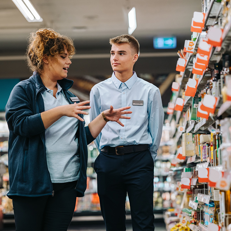 Two coworkers wearing uniforms and nametags talk in front of a shelf full of products