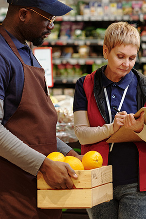 Administrator of supermarket making notes in document while standing by sales assistant with box of fresh oranges in grocery department